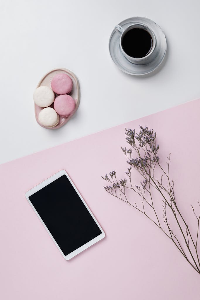 Aesthetic flatlay featuring macarons, a smartphone, coffee, and a dried floral arrangement on a pink background.