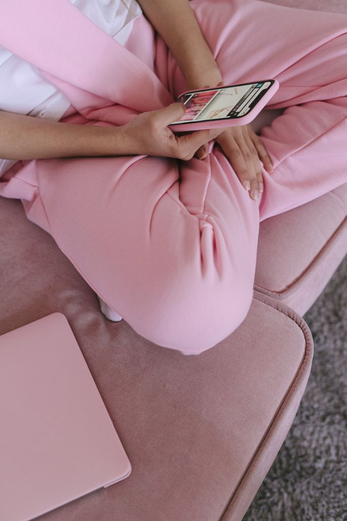 Close-up of a woman using a smartphone on a pink couch with a laptop nearby, creating a cozy and modern tech-savvy vibe.