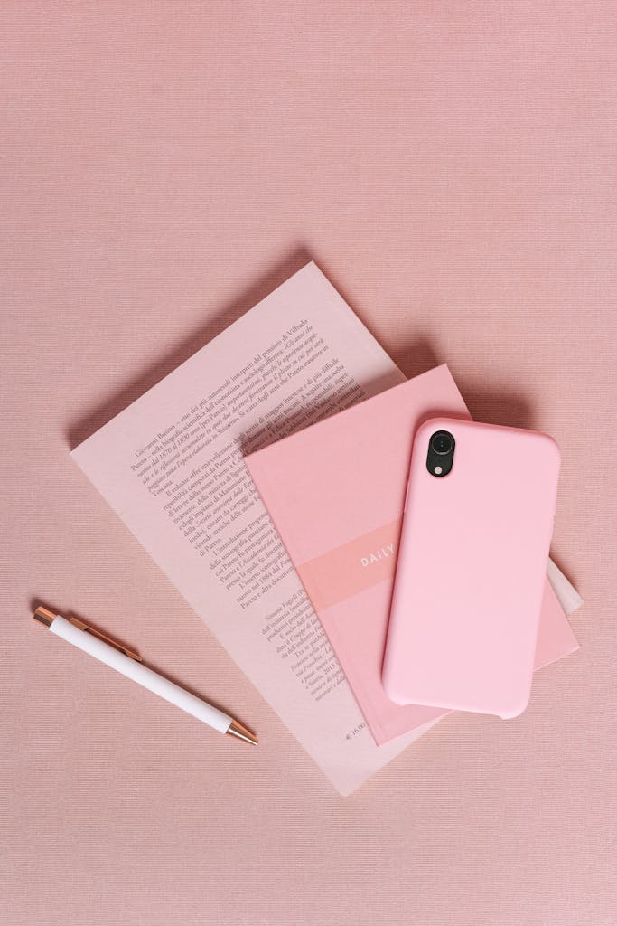 Flat lay of a pink themed workspace featuring a smartphone, notebook, and pen on a pink surface.